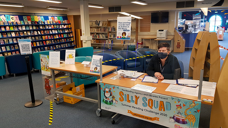 Photo of the inside of Bradley Stoke Library with a member of staff seated behind a screen and wearing a mask.