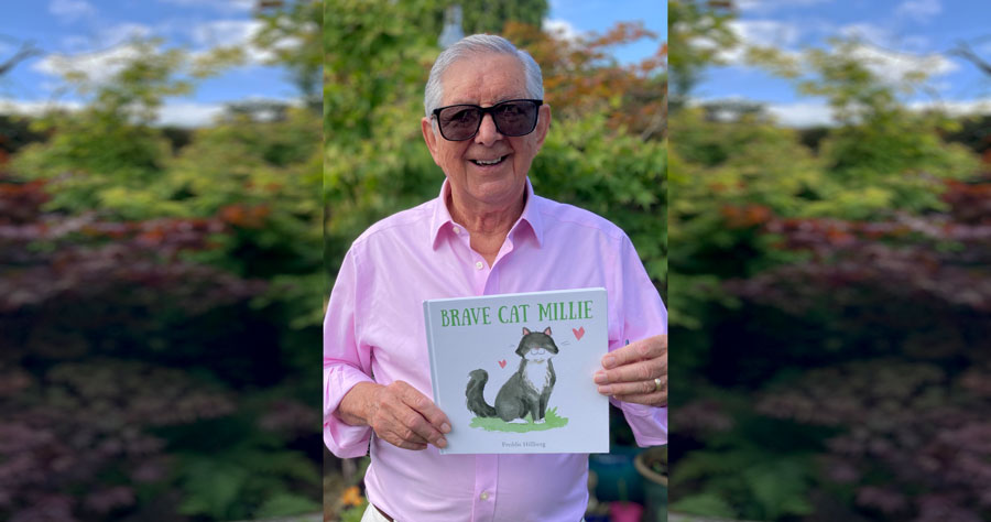 Photo of a man in a pink shirt holding a book that has an illustration of a cat on the cover.